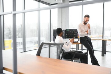 Two diverse colleagues traders talking to each other, sitting in the office in front of multiple computer screens. Stock trading, people, business concept