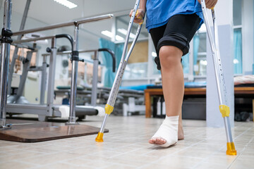 Medical care patient walking using crutches to support her injured ankle sprain, in the hospital physical therapist room.