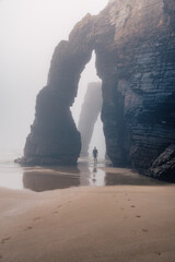 Man standing under natural arch on Cathedrals beach in Galicia, Spainn. Tourist silhouette in foggy landscape with Playa de Las Catedrales Catedrais beach in Ribadeo, Lugo on Cantabrian coast - 784679839