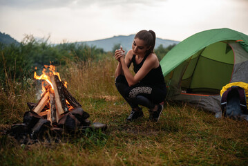 Attractive caucasian woman squatting near green tent and warming hands over bonfire. Active female...