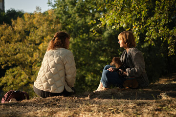Two friends talking and laughing sitting on the grass in a park. Drinking coffee to go, with small dog in the sweater. Sunny evening light, weekend concept. High quality photo