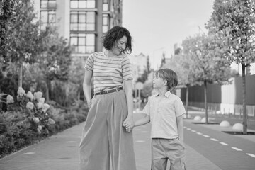 Obraz premium Caucasian young beautiful happy mother and cute small son walking the street and looking at each other. Woman and little boy smiling, talking and having vacations. High quality photo