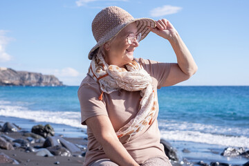 Smiling senior Caucasian woman on seaside vacation holding her hat so it doesn't fly away while...