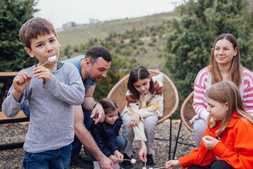 Big happy family roasting mashmallows outdoors. Little cute boy eating hot marshmallow on stick.