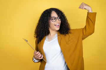 African american business woman with paperwork in hands over yellow background showing arms muscles smiling proud