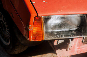 The front of a red old retro USSR car with peeling paint with a headlight without a bumper after an accident.