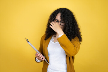 African american business woman with paperwork in hands over yellow background smelling something stinky and disgusting, intolerable smell, holding breath with fingers on nose - obrazy, fototapety, plakaty