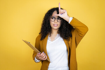 African american business woman with paperwork in hands over yellow background making fun of people with fingers on forehead doing loser gesture mocking and insulting.