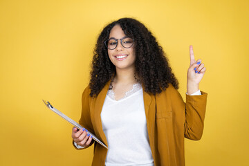 African american business woman with paperwork in hands over yellow background smiling, looking at the camera and pointing up with fingers and raised arms