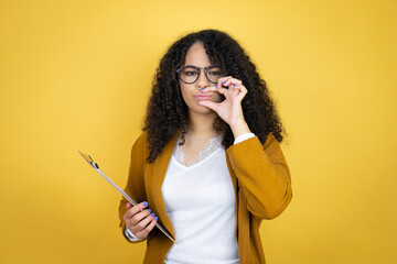 African american business woman with paperwork in hands over yellow background mouth and lips shut as zip with fingers. Secret and silent, taboo talking