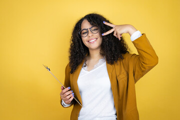 African american business woman with paperwork in hands over yellow background Doing peace symbol with fingers over face, smiling cheerful showing victory