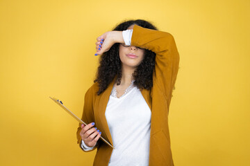 African american business woman with paperwork in hands over yellow background covering eyes with arm smiling cheerful and funny. Blind concept.