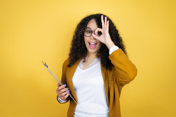 African american business woman with paperwork in hands over yellow background doing ok gesture shocked with smiling face, eye looking through fingers