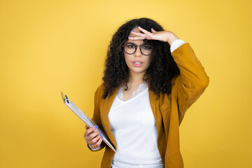 African american business woman with paperwork in hands over yellow background very happy and smiling looking far away with hand over head. searching concept.
