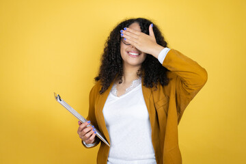African american business woman with paperwork in hands over yellow background covering eyes with hands smiling cheerful and funny. Blind concept.