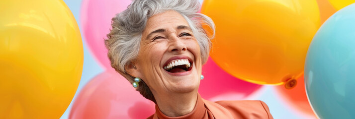 An elderly woman with gray hair is laughing heartily, dressed in orange and surrounded by vibrant balloons, radiating happiness