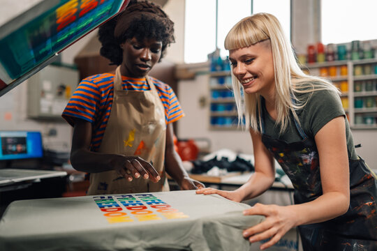 Happy interracial female print shop workers silkscreen printing on machine