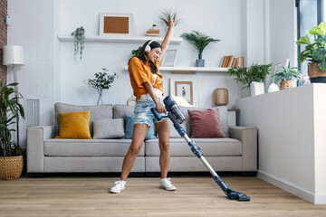 Shot of young happy woman listening and dancing to music while cleaning the living room floor with a vaccum cleaner - obrazy, fototapety, plakaty
