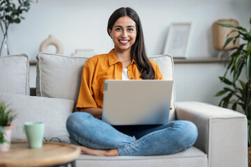 Smiling young woman working with her laptop while sitting on a couch at home