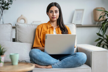 Serious young woman working with her laptop while looking at camera sitting on couch at home.