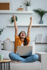 Young happy woman celebrating something while working with laptop sitting on a couch at home
