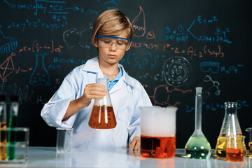 Blonde hair schoolboy in laboratory wear lab coat stand and learn science of chemistry technology in STEM class. The student hold beaker with orange liquid. On the table put many flask. Erudition.