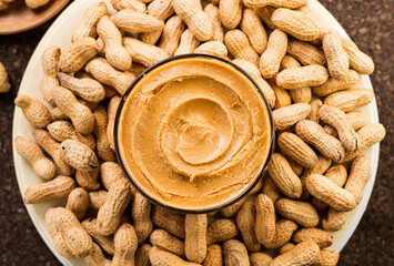 Peanut butter in a glass bowl over raw peanuts background. Creamy smooth peanut butter in glass bowl backdrop. Texture. Organic food. American cuisine. Top view