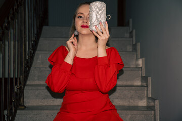 glamourous young beautiful woman in red dress, with silver bag and silver earrings is sitting on...