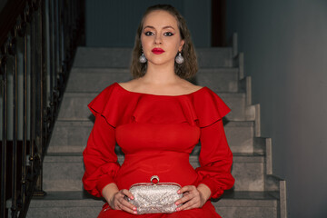 glamourous young beautiful woman in red dress, with silver bag and silver earrings is sitting on the staircase, vintage retro style