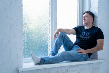 young handsome man with sad face, wearing jeans and t shirt is sitting on windowsill near the window, man looks through the window and thinks about life 
