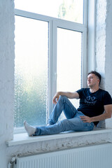 young handsome man with sad face, wearing jeans and t shirt is sitting on windowsill near the window, man looks through the window and thinks about life 