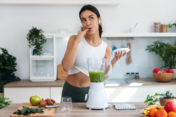 Athletic woman preparing smothie while listening music with earphones in the kitchen at home.