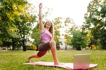 Young woman follows with a laptop a fit exercises  in the park. Online training, course. Training...