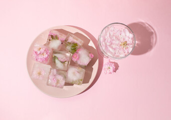 Beautiful small pink flowers in ice cubes and glass with petals on pink plate on pink background, top view