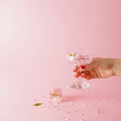 A woman's hand holds a glass with rose petals, an ice cube is on the table on a pink background
