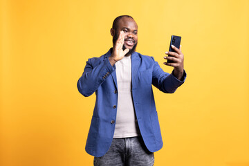 Joyful african american man greeting friends during teleconference meeting using smartphone, studio...