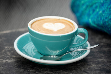 Cup of cappuccino with a heart shaped foam design in a turquoise cup, on a wooden table with a blue sweater in the background.