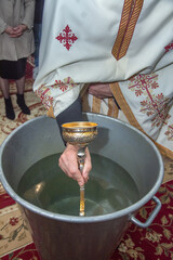 an orthodox priest inserts a cross into the metal baptismal vessel