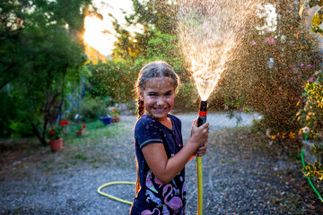 A joyful girl plays with a garden hose, spraying water in the golden light of the setting sun,...