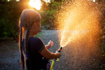 A joyful girl plays with a garden hose, spraying water in the golden light of the setting sun, creating a misty spray against a background of trees.