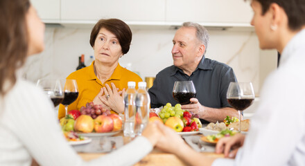 Worried elderly mother sitting at cozy set table with husband during heartfelt conversation with young adult daughter and son-in-law at family dinner
