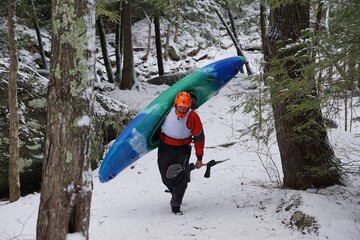 Man carrying blue and green kayak in the snow, wearing an orange helmet and carrying a paddle