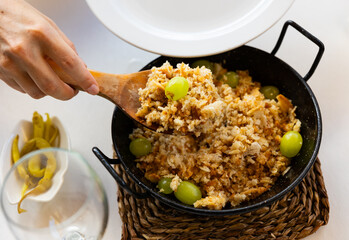 Female hands ladling Migas del pastor, fried bread crumbs with fresh grapes and pickled piparra peppers into plate. Culinary traditions of Aragon region in Spain - obrazy, fototapety, plakaty