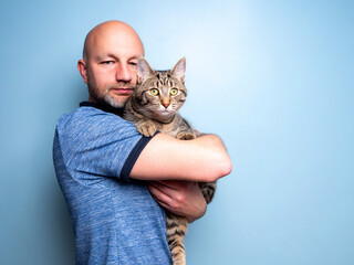 Man holding cute tabby cat in his hands blue wall background. Display of love and care towards home pet. Male model is bold with short beard in blue shirt. Expression of sweet emotions and protection
