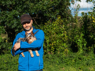 Man in blue jacket holding in his hands cute small Yorkshire terrier, green trees and sky...