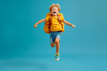 Obraz premium Cheerful smiling little boy with big school backpack isolated on blue empty background with space for text or inscriptions, back to school theme 