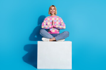 Full length photo of focused appreciative girl wear colorful pullover sit on platform in meditation pose isolated on blue color background