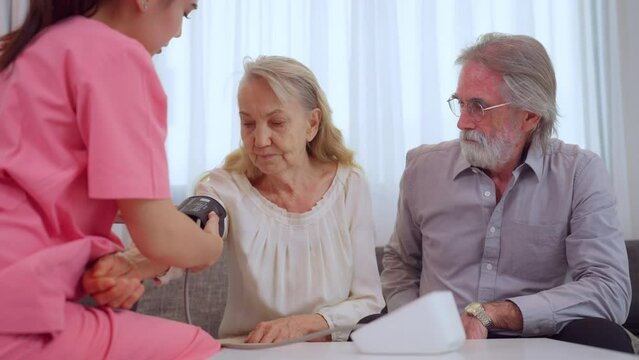 Elderly woman having her blood pressure taken by a nurse while senior man watches with concern.