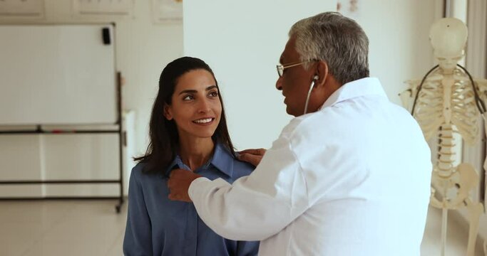 Elderly Indian doctor man listening to heartbeat rate of young patient, applying stethoscope to chest. Positive young Hispanic woman asking practitioner questions on checkup meeting