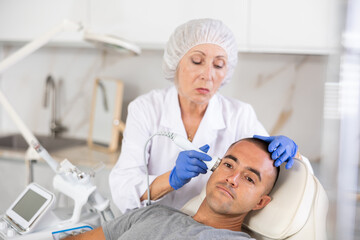 Female cosmetologist performing anti-aging face phototherapy procedure for male client in clinic of aesthetic medicine